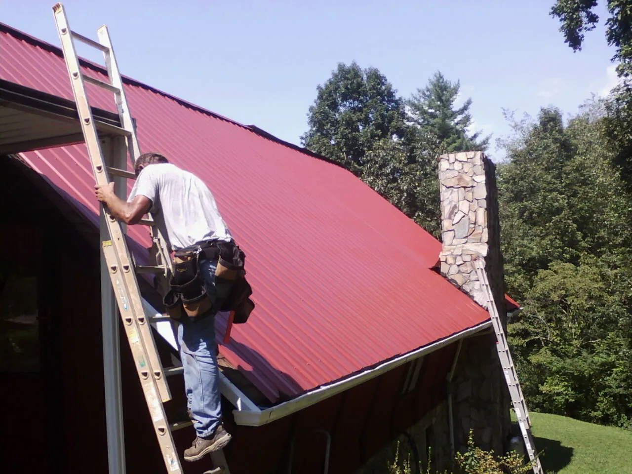 Roofer on a ladder beside a red metal roof and stone chimney — Soffit & Fascia Repair by Roofing Brothers in Cleaton